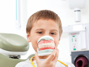 Ein Junge im Behandlungsstuhl eines Zahnarztes hält sich ein künstliches Gebiss vor das Gesicht