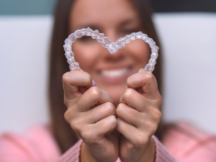 Eine junge Frau hält zwei unsichtbare Zahnschienen so in die Höhe, dass sie sich an zwei Enden berühren und die Form eines Herzens ergeben