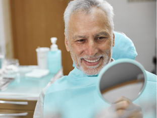 Älterer Mann mit grauen Haaren und Bart sitzt auf dem Zahnarztstuhl und lächelt in einen Handspiegel
