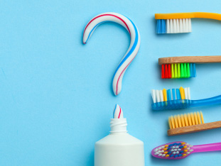 Eine anzahl verschiedener, bunter Zahnbürsten liegt neben einem mit Zahnpasta geschriebenen Fragezeichen