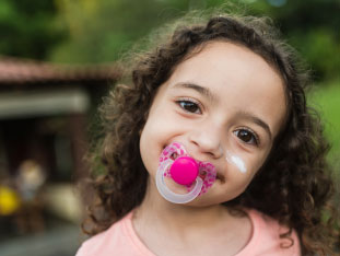 Ein kleines, etwa dreijähriges Mädchen mit  Schnuller im Mund lächelt in die Kamera